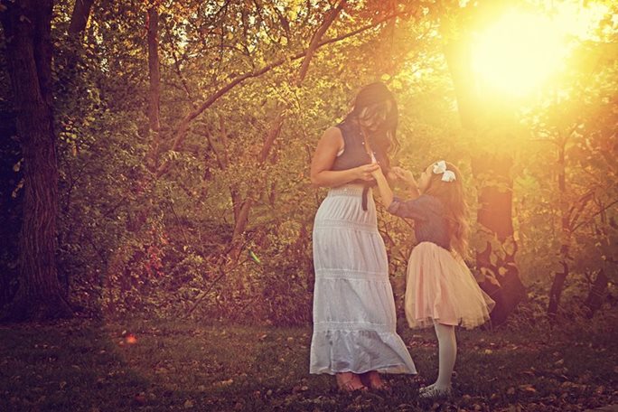 A young girl looks up at her mother in a wooded setting with the setting sun casting a golden light through the trees