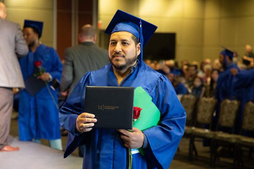 Israel Valdez holding his MCC diploma wearing graduation cap and gown