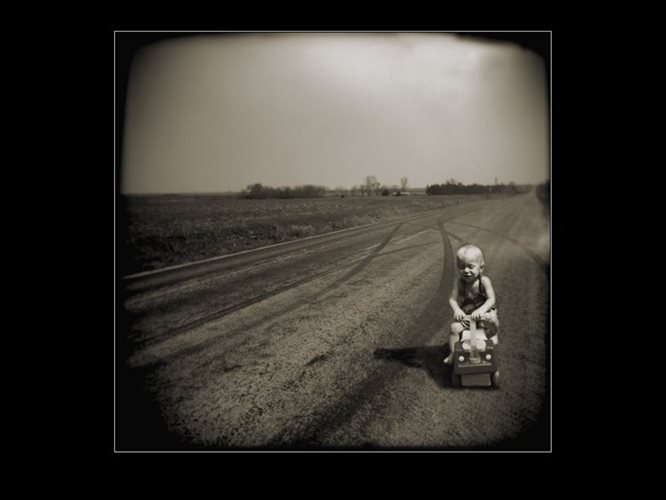 Black and white photograph of a young boy riding a toy car on a dirt road. There is an empty field in the background, and the sky is large and clear.