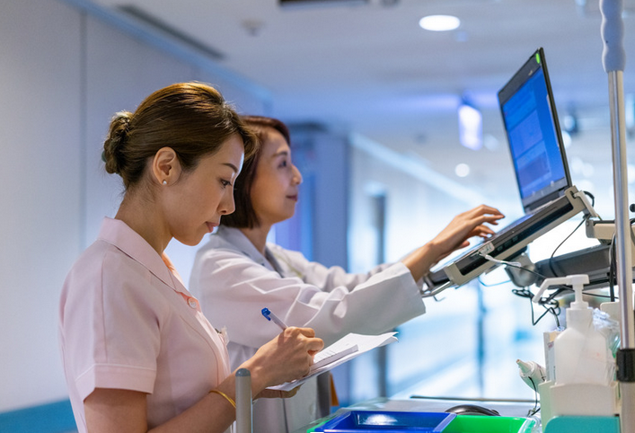 one medical scribe working at a computer while the other checks writes information on a printout