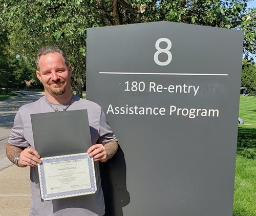 A man shows off his degree in front of the 180 Re-entry Assistance Program building sign