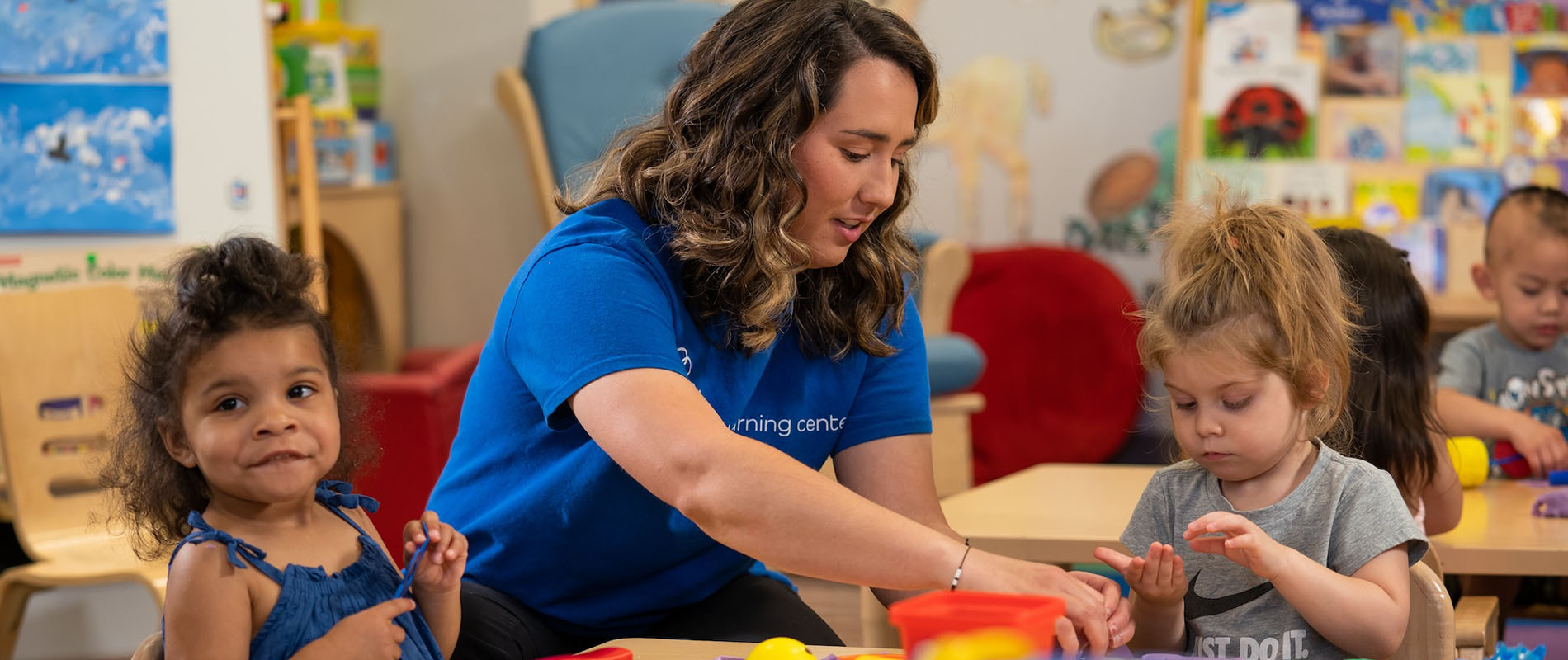 A woman early childhood education professional works assists two young girls sitting at a table engaged in a group classroom project in a childcare setting.